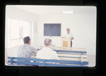 man standing behind a pulpit inside a church building or classroom by Haven L. Miller
